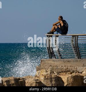 Tel Aviv, Israel - 15. September 2022: Ein junger Mann sitzt auf dem Geländer der Uferpromenade von Tel Aviv und sieht zu, wie die mediterranen Wellen auf dem Shor brechen Stockfoto