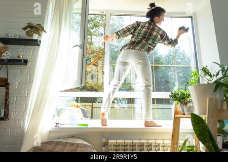 Frau wäscht das Fenster des Hauses manuell mit einem Lappen mit Reinigungsspray und Mopp innen mit weißen Vorhängen. Ordnung und Sauberkeit wiederherstellen Stockfoto