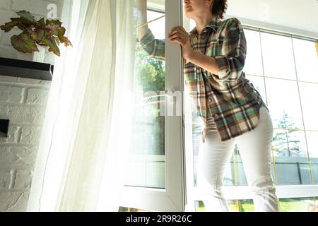 Frau wäscht das Fenster des Hauses manuell mit einem Lappen mit Reinigungsspray und Mopp innen mit weißen Vorhängen. Ordnung und Sauberkeit wiederherstellen Stockfoto