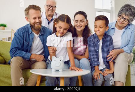 Das Mädchen legt eine Münze in die weiße Sparschweinbank, während die ganze Familie sie zu Hause anschaut. Stockfoto