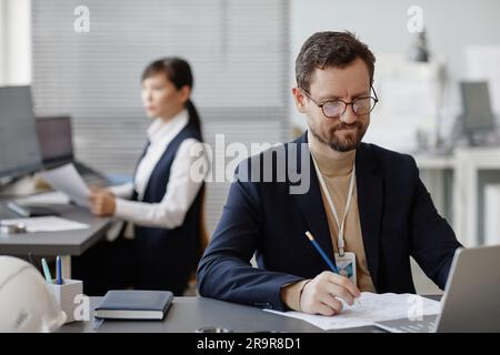 Porträt eines bärtigen männlichen Ingenieurs, der Computer mit Bauplänen und Plänen benutzt, während er Berechnungen im Büro und im Kopierraum macht Stockfoto