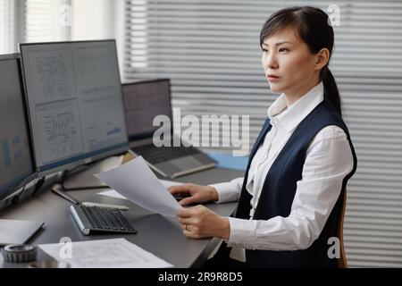 Seitenansicht einer Asiatin als Ingenieurin, die Computer mit Bauplänen und Plänen benutzt, während sie im Büro rechnet Stockfoto