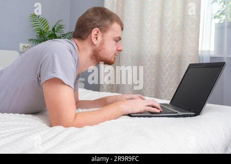 Ein Mann mit Laptop liegt im Bett. Freiberufler arbeiten von zu Hause aus. Fernunterricht. Online-Shopping ohne das Haus zu verlassen. Surfen im Internet. Spielsucht. Black-Friday-Angebot. Cyber Monday. Stockfoto