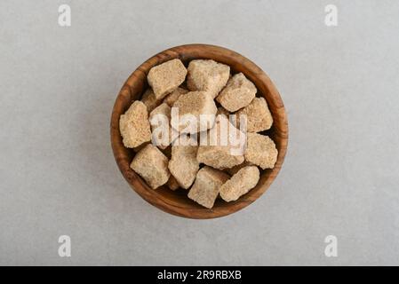 Braune Rohrzuckerwürfel in Holzschüssel auf Betonhintergrund. Draufsicht Stockfoto