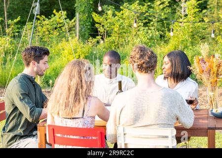 Gartenfreunde: Multiethnische Freunde vereinen sich am Abend. Hochwertige Fotos, erleben Sie den Zauber einer abendlichen Zusammenkunft in einem wunderschönen Garten, wo eine Gruppe von Freunden aus verschiedenen Rassen zu einem köstlichen Abendessen und Momenten der reinen Freude zusammenkommt. Dieses fesselnde Foto zeigt die lebendige Energie und warme Kameradschaft, während sie an einem Tisch sitzen und Lachen, Geschichten und ein köstliches Essen teilen. Die Gartenanlage verleiht der Szene einen Hauch von natürlicher Pracht und schafft die perfekte Kulisse für ihre lebhaften Gespräche und wertvollen Erinnerungen. Treten Sie dieser vielfältigen Gruppe bei Stockfoto