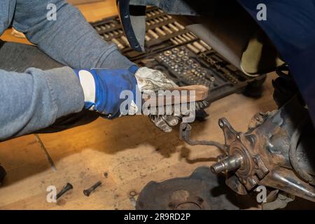 Der Automechaniker reinigt die Bremsleitung mit einer Drahtbürste. Löst und wartet das Radnabenlager. Kit-Box im Hintergrund. Stockfoto