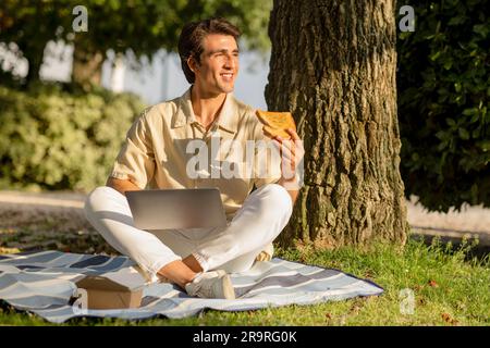 Ein glücklicher Mann macht ein Picknick im Park, benutzt einen Laptop, isst Sandwich Stockfoto
