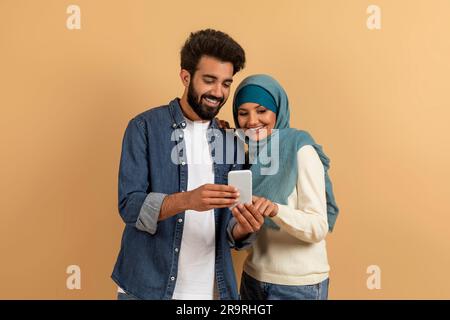 Coole App. Aufgeregtes arabisches Paar, das ein Smartphone zusammen über beigefarbenen Studiohintergrund benutzt Stockfoto