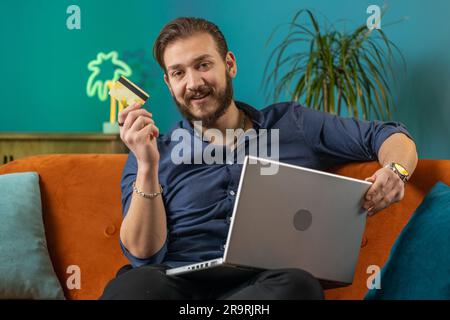 Ein junger libanesischer Mann, der eine Kreditkarte und einen Laptop benutzt, um Geld zu überweisen, online einzukaufen, Essen zu bestellen. Porträt eines arabers im Wohnzimmer auf der Couch Stockfoto