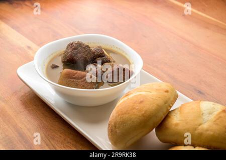 Nahaufnahme einer Schüssel mit dampfender heißer Suppe und ein Stück knusprigem Brot, serviert auf einem weißen Teller Stockfoto