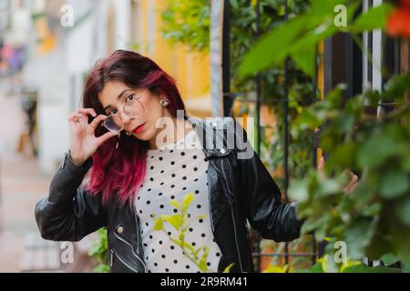 Eine Frau im lässigen Street-Stil, die ein gepunktetes Hemd und eine schwarze Lederjacke trägt und mit ihrem Handy spricht, während sie in einer Stadtumgebung steht Stockfoto