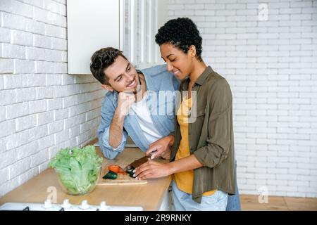 Positives, modernes, multiethnisches Paar, verliebt, in lässiger Kleidung, junge Ehepartner, stehen zu Hause in der Küche, bereiten Salat zu, Frau schneidet Gemüse, sie sind froh, Zeit miteinander zu verbringen, lächeln Stockfoto