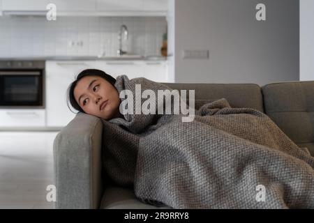 Frustrierte asiatische Frau, die auf der Couch liegt, sieht leider durch Depressionen gewickelt in Karo zu Hause aus. Stockfoto