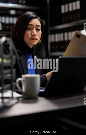 Ein asiatischer Privatdetektiv, der die Details des Strafverfahrens auf dem Laptop überprüft, bevor er mit der Untersuchung beginnt. Detective im Archivraum mit Kriminologie-Akten auf Schrankregalen Stockfoto