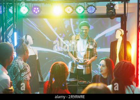 Leute tanzen und genießen dj-Show auf Disco-Party im Nachtclub. Junger Mann, Musiker, der elektronische Musik auf der Bühne spielt, für die Menge im Club, beleuchtet mit Lichtern Stockfoto