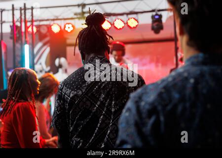 Verschiedene Clubmitglieder feiern auf Tanzfläche, während sie ein elektronisches Musikkonzert im Nachtclub besuchen. Die Leute tanzen in Diskotheken, während dj auf der Bühne im Club auftritt Stockfoto