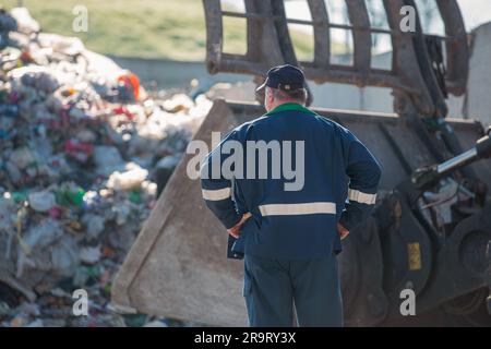 Ein Arbeiter im Recycling-Center, in dunkelblauer Arbeitskleidung, der sich einen unsortierten Müllhaufen ansieht, ist gerade in der Materialrückgewinnungsanlage angekommen, Rückansicht. Stockfoto