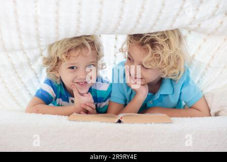 Lesebuch für Kinder im Bett unter der Strickdecke. Zwei Brüder spielen zusammen. Kinder gemütliches Zimmer im Hyggge-Stil. Kleiner Junge, der Hausaufgaben macht, bevor er schläft. Stockfoto