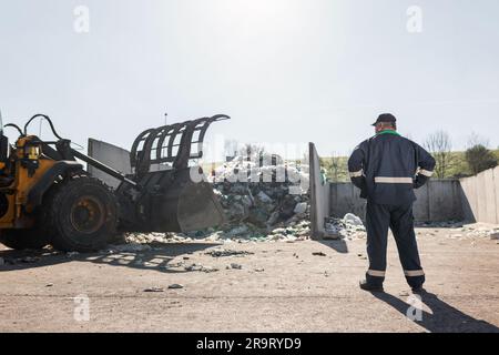 Ein Mitarbeiter des Recyclingzentrums in dunkelblauer Arbeitskleidung, der sich einen unsortierten Müllhaufen ansieht, ist gerade in der Materialrückgewinnungsanlage eingetroffen Stockfoto