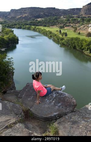 Eine Frau in einem rosa Hemd macht eine Pause von der Wanderung und sitzt auf einem großen Felsen mit Blick auf den Snake River in Twin Falls, Idaho. Stockfoto
