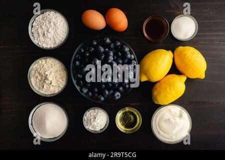 Blaubeermuffins mit Zitronenglasur auf dunklem Waldhintergrund: Frische Blaubeeren, Zitronen und andere Rohstoffe auf rustikalem Holzhintergrund Stockfoto