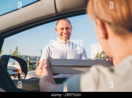 Ein junger, lächelnder Zusteller, der zwei Pizzakartons an eine Frau übergab, die auf dem Autositz sitzt, an die Straße der Stadt. Kleine Unternehmen, Menschen-Relatio Stockfoto