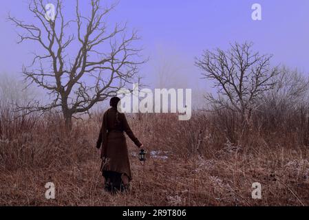 Viktorianische Frau, die mit Laterne durch das Wintermarsch wandert. Der frühe Nebel ruft eine andere Welt und eine andere Zeit hervor. Stockfoto