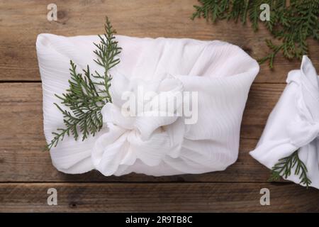 Furoshiki-Technik. Geschenke verpackt in weißem Stoff und Thuja-Äste auf Holztisch, flach liegend Stockfoto