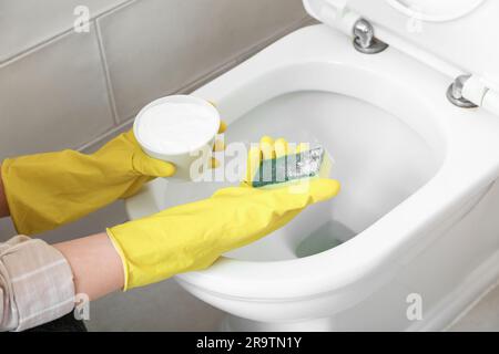 Frau in Gummihandschuhen, die weiße Toilettenschüssel mit Backpulver und Schwamm reinigt Stockfoto