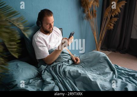 Ein Mann mit Bart, der im Bett liegt und Kopfhörer trägt, sieht sich das Smartphone an und hört Lieblingsmusik. Entspannen Sie sich allein zu Hause Stockfoto