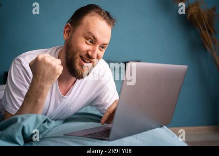 Porträt eines glücklichen Mannes, der sein Wochenende im Bett verbringt, Fußball live auf seinem Laptop spielt und Geld mit Wetten auf ihn feiert Stockfoto