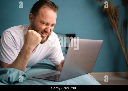 Aufgeregter, überraschter junger Mann, der ja-Gesten macht, auf den Laptop-Bildschirm schaut, sich nach dem Aufwachen im Bett entspannt. Guy erhielt eine E-Mail, in der er Online-Lotterie feierte Stockfoto