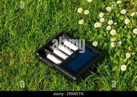 Schwarzes AA-Batterieladegerät mit wiederaufladbaren Alkali-Batterien auf dem Rasen. Der Akku wird geladen. Stockfoto