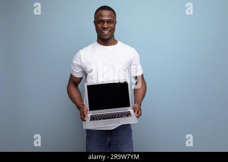 Erfolgreicher junger afrikanischer Mann im weißen T-Shirt, der per Fernzugriff mit einem Laptop im Studiohintergrund und Kopierbereich arbeitet Stockfoto