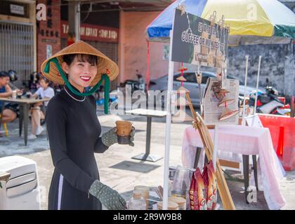 Februar 25 2023 - George Town - Penang - Malaysia - wunderschöne junge Frau aus Malaysia mit einem speziellen Hut und schwarz gekleidet verkauft Kaffee an Touristen smi Stockfoto