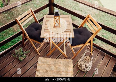 Moderner, kleiner Holzbalkon mit Sitzecke, Gartenmöbeln aus Holz, weichen schwarzen Kissen, Laterne mit brennender Kerze auf dem Tisch im Freien. Stockfoto