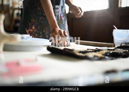 Handwerkerin in Schürze knete ein Stück rohen Ton mit den Händen auf einem Holztisch. Handwerk, Kreativität, Hobby und Aktivitätskonzept Stockfoto