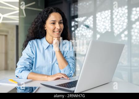 Fröhliche und lächelnde hispanische Geschäftsfrau, die auf dem Laptop tippt, Büroangestellter mit lockigem Haar, zufrieden mit den Ergebnissen, bei der Arbeit im Bürogebäude Stockfoto