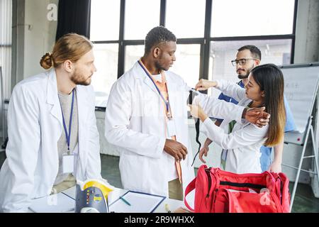 asiatische Frau in weißem Mantel, die Druckstaub auf den Arm eines afroamerikanischen Studenten in der Nähe der Lehrerin aufträgt, medizinische Ausrüstung und erste-Hilfe-Kasten, lif Stockfoto