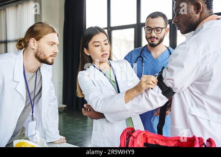 Sanitäter in der Brille, der eine junge asiatische Frau ansieht, die während des erste-Hilfe-Seminars in tr einen Kompressionsstrumpf am Arm eines afroamerikanischen Schülers anlegt Stockfoto