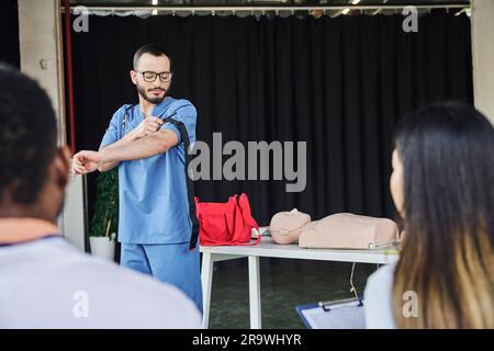 Erste-Hilfe-Seminar, medizinischer Lehrer in blauer Uniform und Brille mit Kompressionsstrumpf in der Nähe von interrassistischen Teilnehmern auf verschwommenem Vordergrund Stockfoto