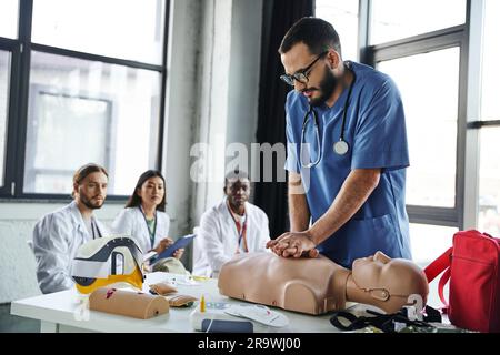 Junge multikulturelle Studenten in weißen Mänteln, die einen medizinischen Lehrer betrachten, der Thoraxkompressionen auf der HLW-Prüfpuppe während des erste-Hilfe-Seminars, Acquiring durchführt Stockfoto