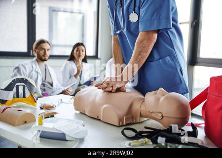 Professioneller Sanitäter in blauer Uniform, der Thoraxkompressionen auf der HLW-Prüfpuppe in der Nähe von medizinischen Geräten und junge multiethnische Studenten auf verschwommenem Rücken Stockfoto