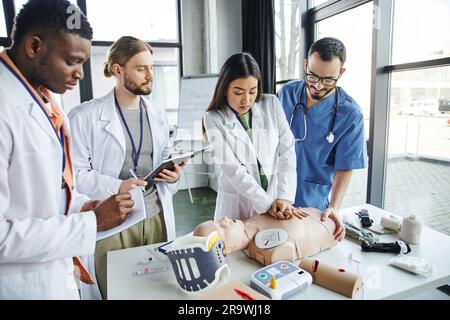 Junge asiatische Frau in weißem Mantel, die Brustkompressionen auf der HLW-Puppe in der Nähe von Lehrer, medizinischen Geräten und multikulturellen Schülern mit Notizbüchern macht, Stockfoto