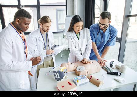 Erste-Hilfe-Seminar, multiethnische Studenten in weißen Kitteln schreiben nahe asiatische Frau, die Thoraxkompressionen auf der HLW-Prüfpuppe mit Hilfe von medizinischem Unterricht Stockfoto