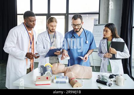 Herz-Wiederbelebung, medizinischer Lehrer hält Defibrillator-Pads über HLW-Übungspuppe in der Nähe von jungen multiethnischen Studenten in weißen Mänteln, Notfallsituationen Stockfoto