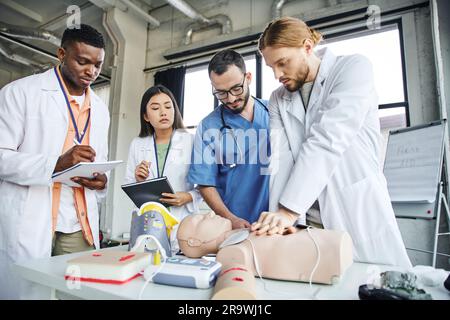 Junger Mann praktiziert Thoraxkompressionen auf HLW-Prüfpuppe in der Nähe von medizinischen Geräten, professionelle Sanitäter und interrassistische Studenten schreiben in Notizbüchern, e Stockfoto