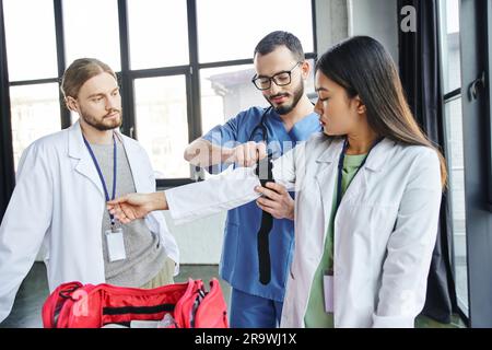 Erste-Hilfe-Lernen, junger Sanitäter mit Brille und Uniform, die Kompressionsstrumpf auf den Arm eines asiatischen Schülers im weißen Kittel, Life-sav anlegt Stockfoto