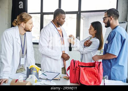 asiatische Frau in weißem Mantel, die Druckstaub auf den Arm eines afroamerikanischen Studenten in der Nähe eines Sanitäters aufträgt, erste-Hilfe-Kasten und medizinische Ausrüstung, Leben Stockfoto