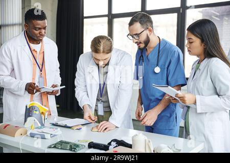 Junge Schülerin in weißer Jacke praktiziert mit Injektions-Trainingspad in der Nähe professioneller Sanitäter und multiethnisches Team mit Notizbüchern in der Nähe medizinischer Ausrüstung Stockfoto